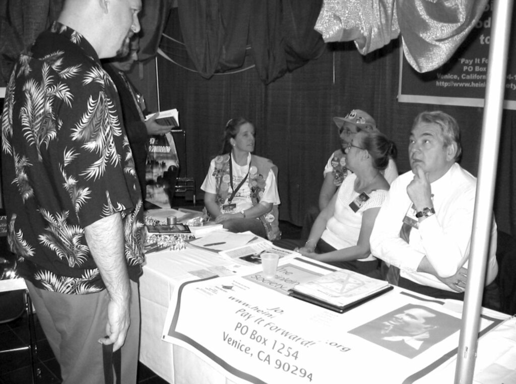Robert James (standing) talks with Bill Patterson (seated) at the Heinlein Society table at Noreascon IV, with Alan Milner, Mike and Sharon Sheffield, Pam Somers in background.